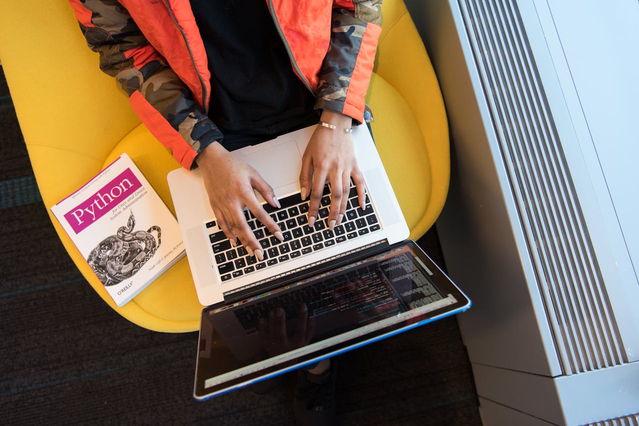 High-angle view of woman coding on a laptop, with a Python book nearby. Ideal for programming and tech content.